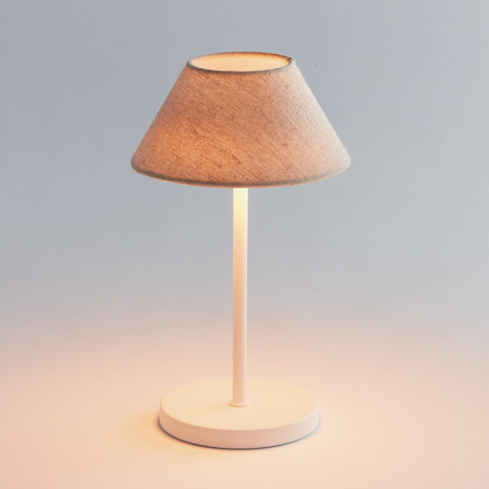 Cordless LED lamp with a beige fabric conical lampshade atop a white cylindrical stem and round base, warm light glow