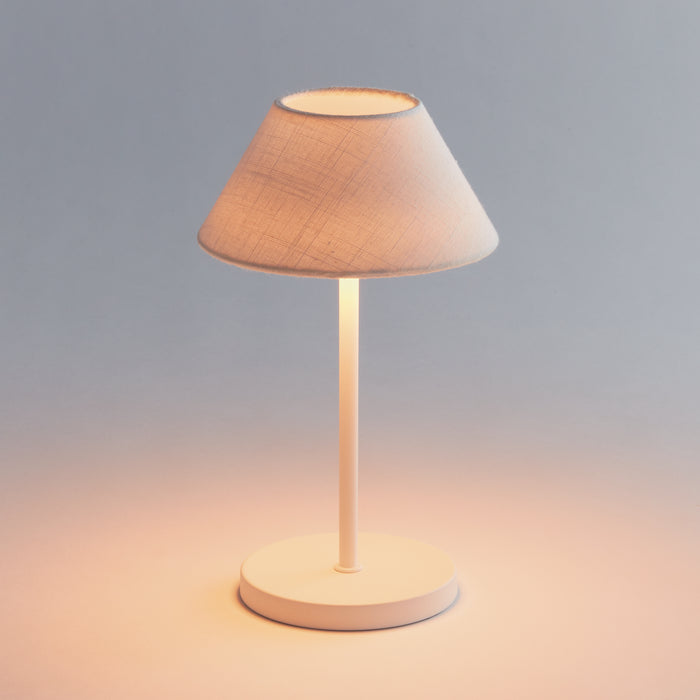 Cordless LED lamp with white metal stand and fabric lampshade, warm glow on a round base, neutral background.