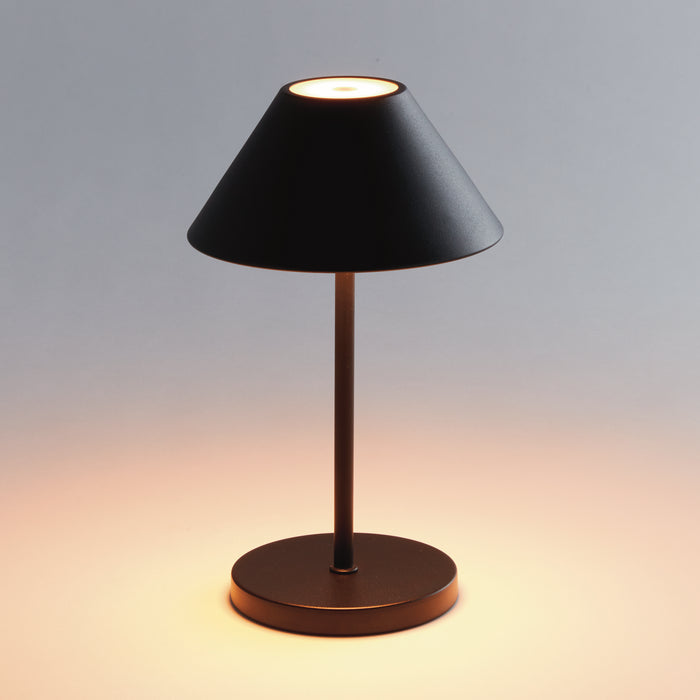Black metal cordless LED table lamp with conical shade, cylindrical stem, round base, glowing warm light on desk.