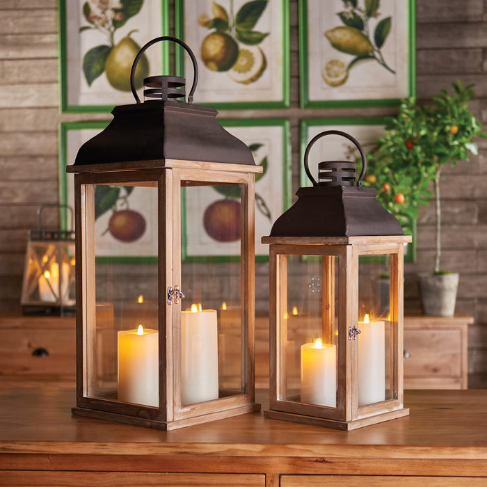 Set of two wooden-framed lanterns with black metal tops, glass panes, and pillar candles inside, on a wooden table.