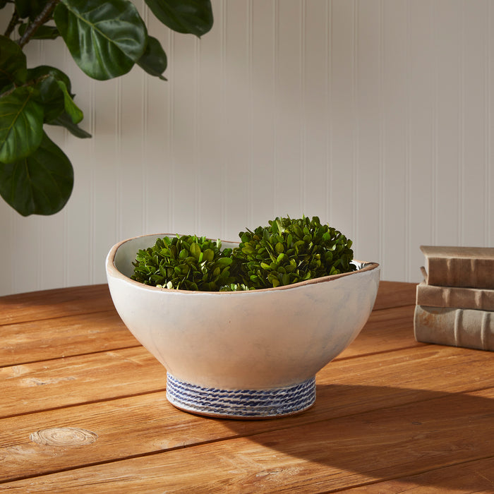 Cream ceramic decorative bowl with blue braided base, containing green trimmed shrubs on a wooden table, neutral background.