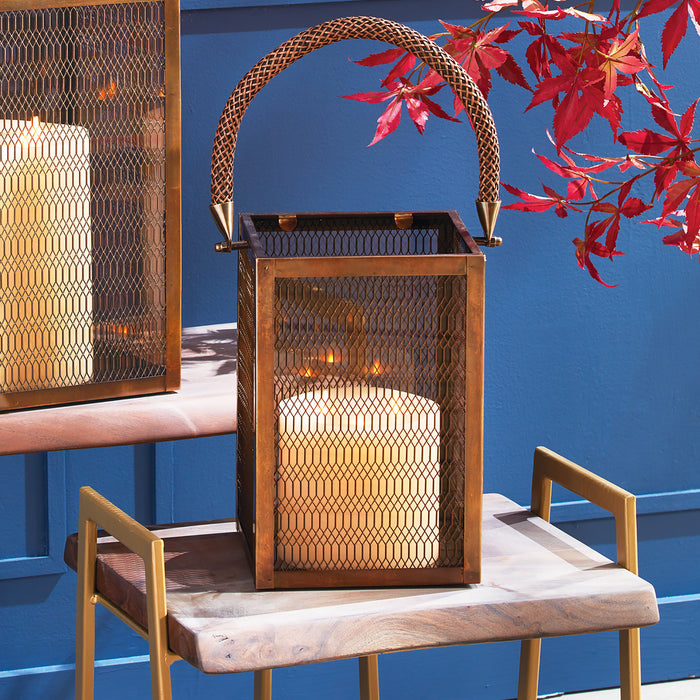 Copper-toned metal lantern with mesh sides and woven arched handle, large cream pillar candle inside, on a wooden table with