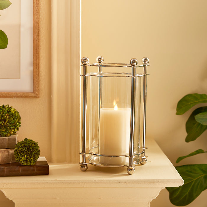 Polished chrome hurricane lantern with a white candle inside on a cream mantel, framed art, green succulents, and leafy
