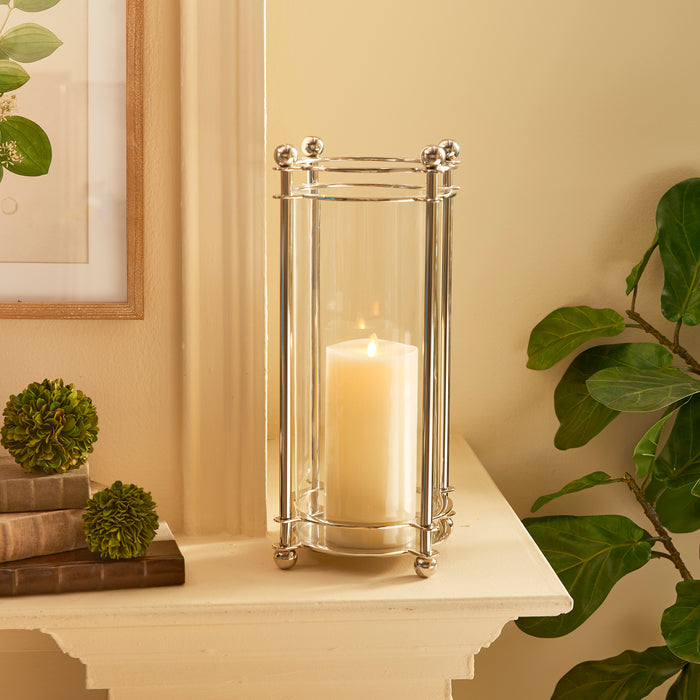 Polished chrome hurricane lantern with a lit ivory candle inside on a white mantel, flanked by greenery.