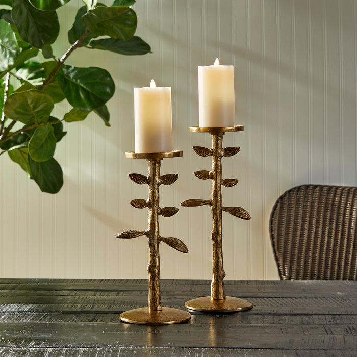 Set of two gold-toned candle stands with textured stems and leaf accents, each holding a white pillar candle; placed on a