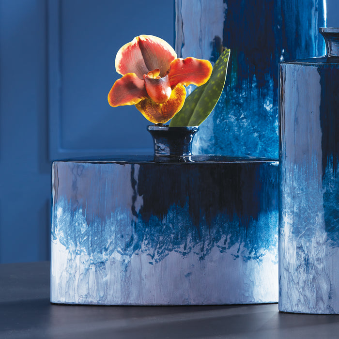 Blue glazed ceramic flask vase with orange orchid stem, set on matching blue marble-textured cylinders, dark backdrop.