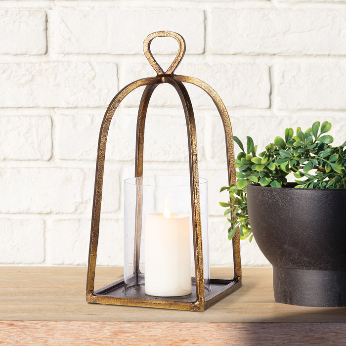 Gold-toned metal lantern with arched frame and loop top, glass cylinder containing white pillar candle; potted greenery on