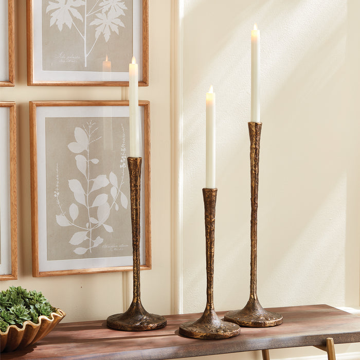 Set of three tall iron taper candle holders with a rustic, textured bronze finish, white candles, on a wooden console with