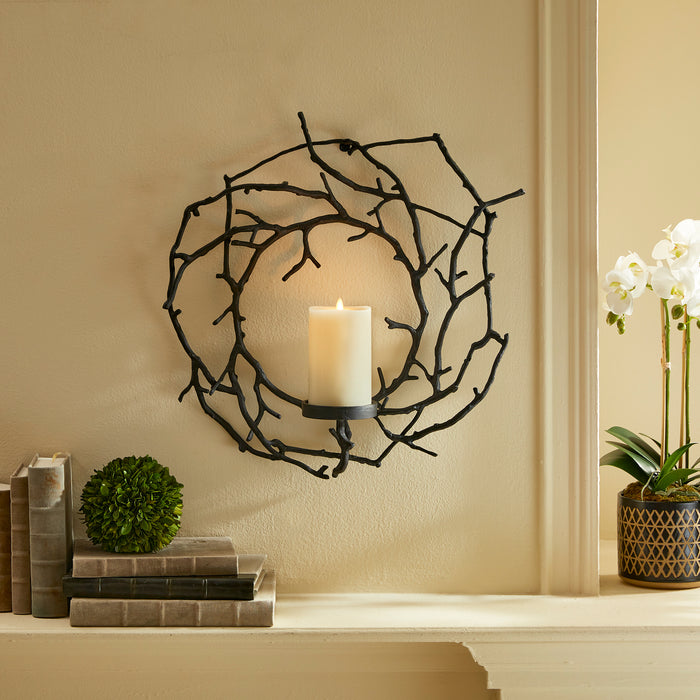 Wrought-iron circular candleholder with intertwining branches holding a lit ivory pillar candle, beige wall background