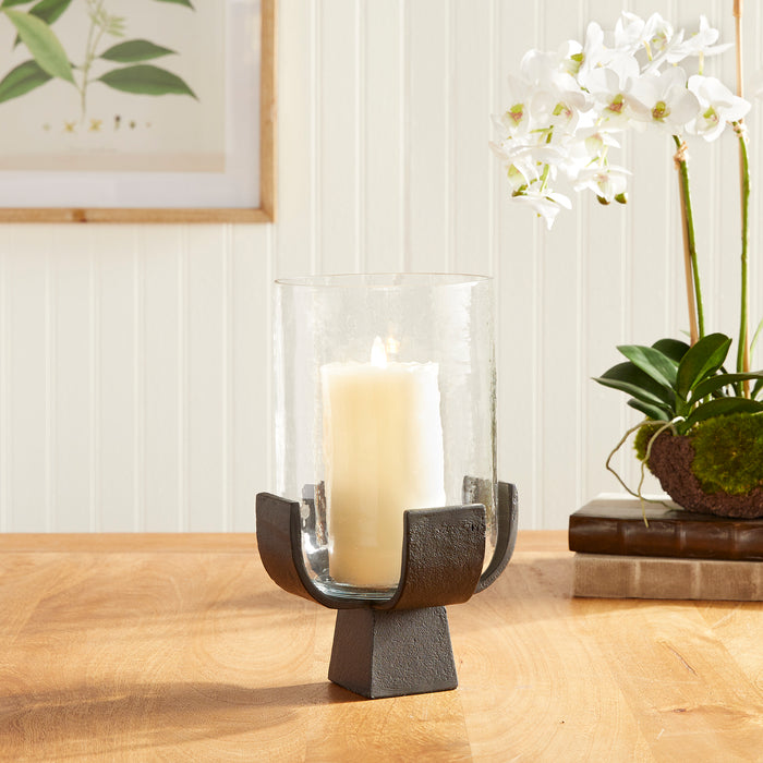 Glass hurricane with a large ivory candle, set in dark metal cradle on a square pedestal base on a wooden table.