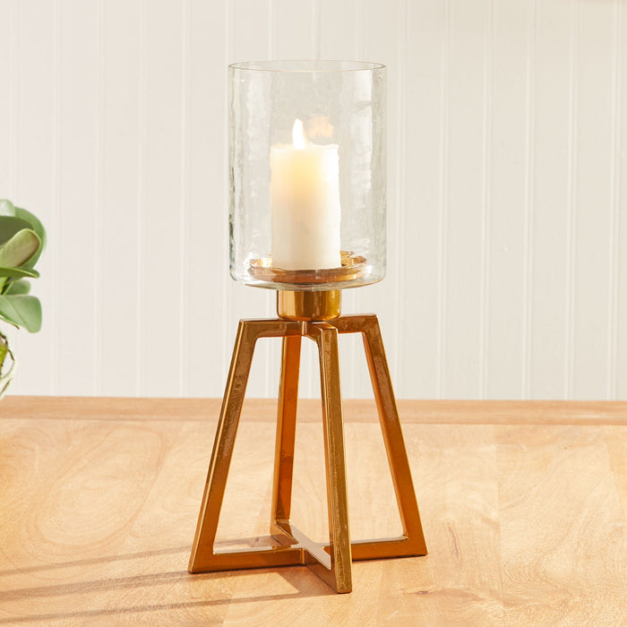 Brass-colored geometric metal candle holder with a glass hurricane and white pillar candle on a wooden surface.