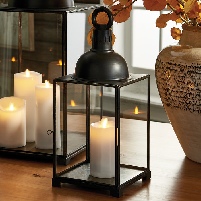 Black metal lantern with glass panels housing a tall white candle, on a wooden table, next to a ceramic vase and lit candles.