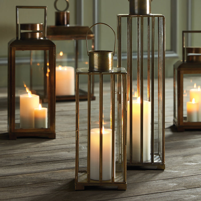 Bronze metal lanterns with glass panes and tall pillar candles on a wooden floor, soft candlelight glow.