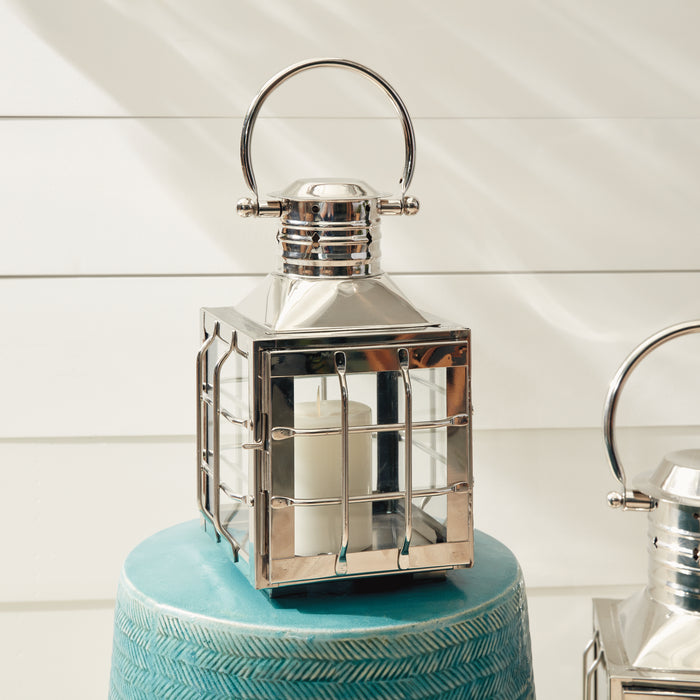 Shiny chrome Nantucket-style outdoor lantern with square cage, glass panes, candle inside, loop handle on top, set on a blue
