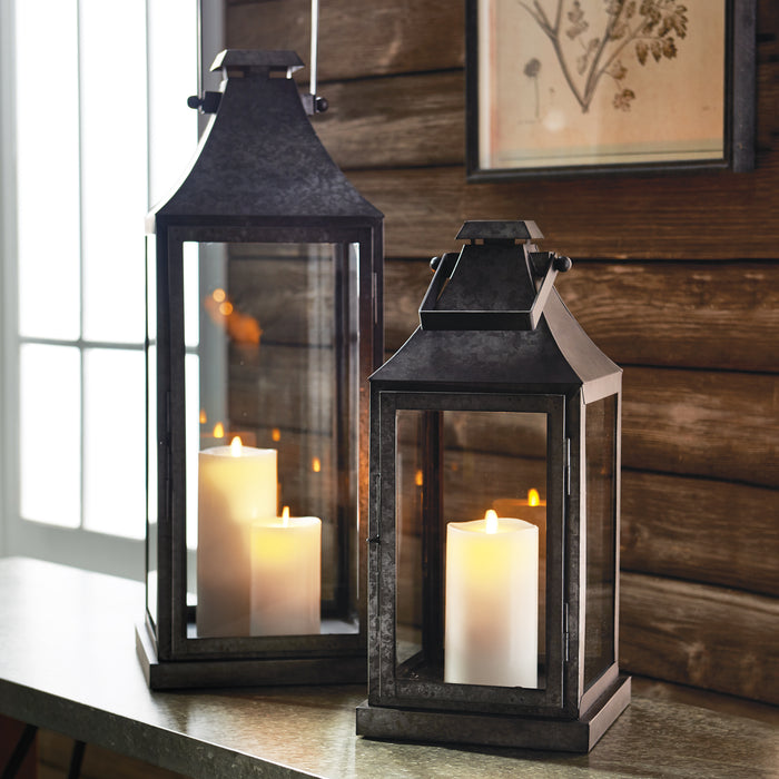Two dark metal rectangular lanterns with glass panes, each containing white candles, set on a wooden surface against a
