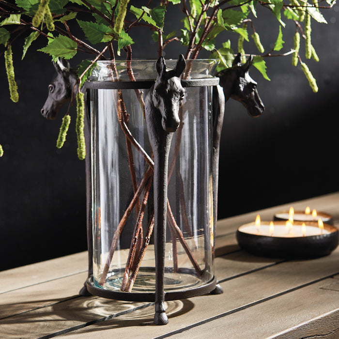 Glass hurricane vase with black horse head iron accents and legs, metal frame, green foliage, on a wooden table with candles