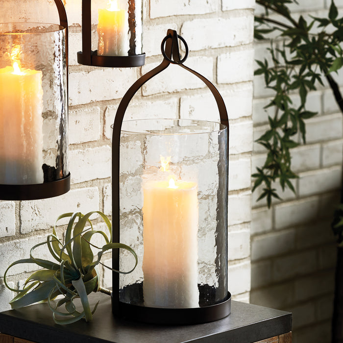 Black metal lantern with tall cylindrical glass shade housing a large white candle, set on a dark base against a white brick