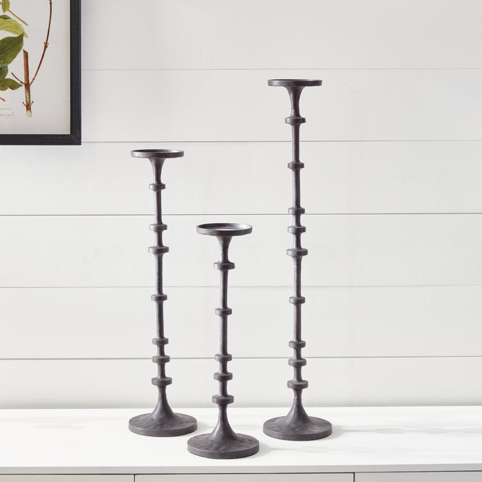 Set of three dark metal taper candle holders with segmented stems on a white surface against a light paneled wall.