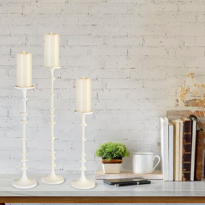 Set of three ivory candle/holder stands with tall, mid, and short heights against a white brick wall, on a marble tabletop
