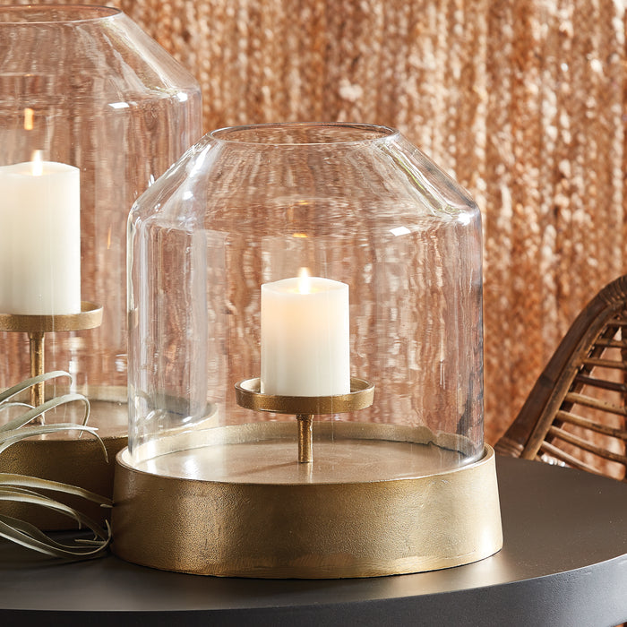 Gold-tone metal hurricane base with a glass dome and a lit white candle on a small pedestal tray, set on a dark surface.