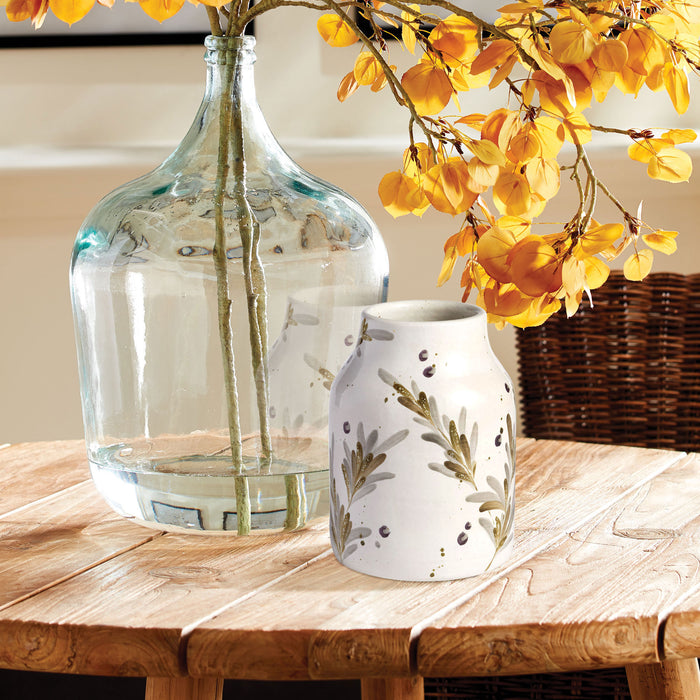 Clear glass vase with yellow autumn branches next to a white ceramic vase with olive branch pattern on a wooden table.