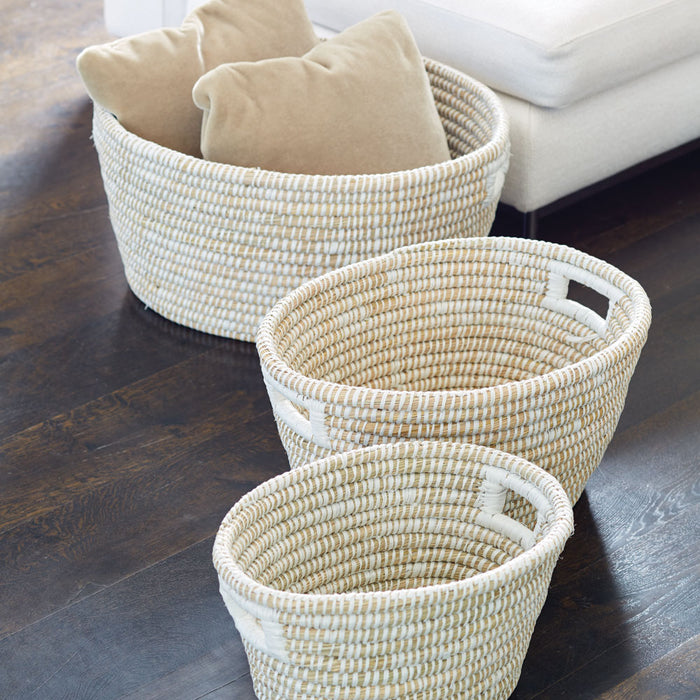 Three woven oval baskets in natural beige with white accents, varying sizes, placed on dark wood floor beside a light sofa.