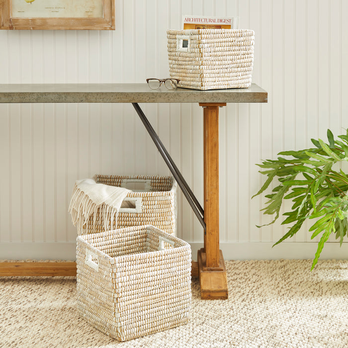 Three handwoven beige and white square basket set with handles, on and beside a wood-legged desk, natural woven texture
