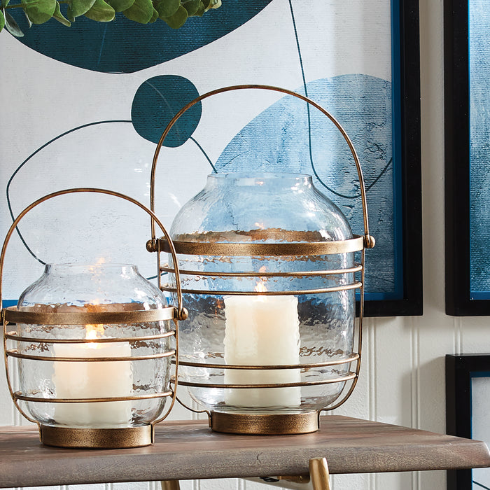 Two glass hurricane lanterns with gold metal frames and handles, candles burning inside, set on a wooden surface with blue
