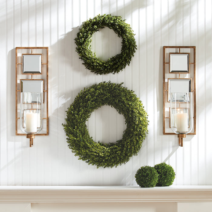 Beige wall with two ornate copper-framed wall sconces flanking two green wreaths; glass candle holders and lit candles.