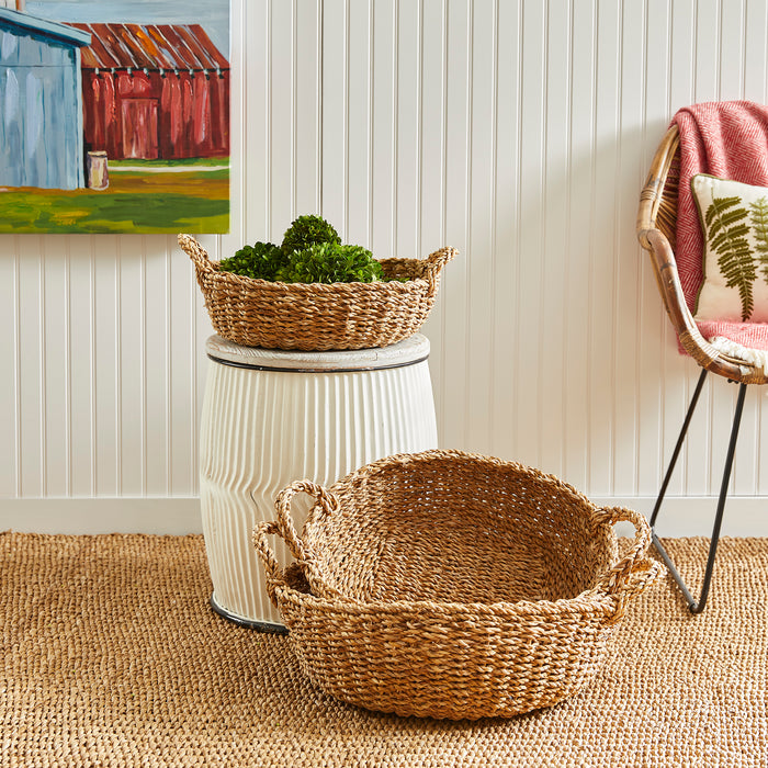 Three seagrass shallow baskets with handles in natural tan, one atop a white ribbed ceramic stool, another empty on a jute