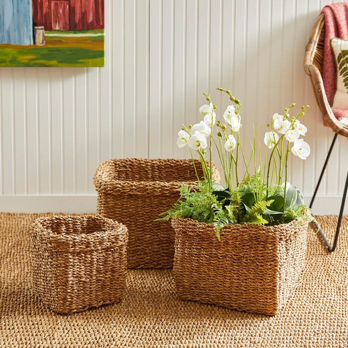 Three seagrass square baskets with cuffs, natural woven texture; largest filled with white orchids and greenery, two empty