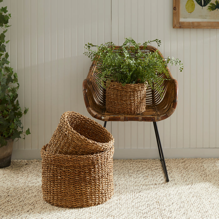 Set of three seagrass round baskets in natural brown tones; one inner basket on chair with greenery spilling over. woven