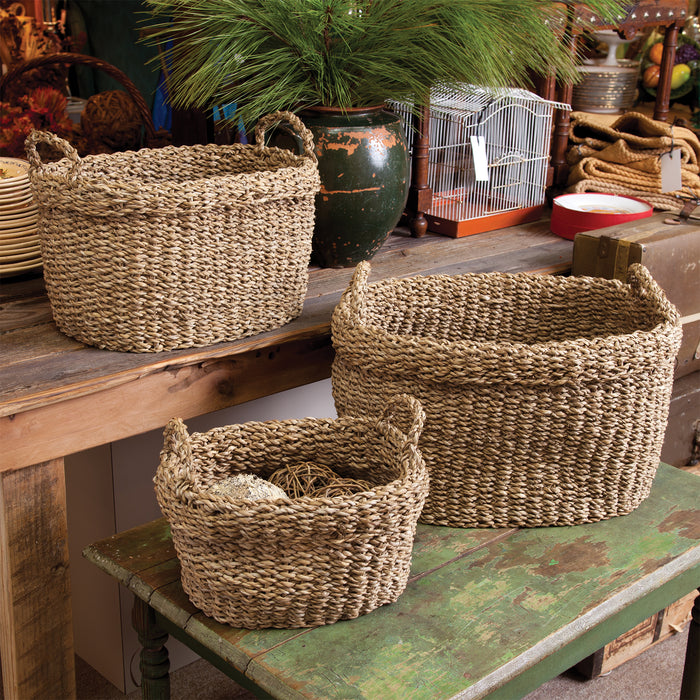 Set of three natural seagrass oval baskets with handles, woven texture, placed on rustic wooden surfaces.
