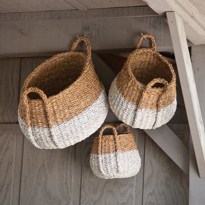 Seagrass round baskets with handles in natural tan and white, set of three in varying sizes, hanging under a wooden porch.