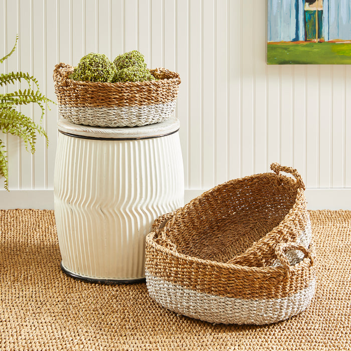 Set of three seagrass baskets with natural tan and light gray bands; two with handles, displayed on woven rug beside a white