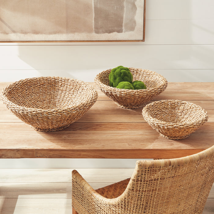 Three seagrass baskets in varying sizes on a wooden table, natural tan color, one holds green moss balls, bright neutral