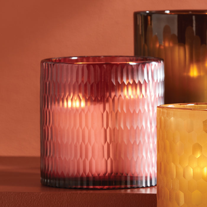 Colorful textured glass hurricane candle holder in pink, with a yellow one to the right and a dark brown behind, on a
