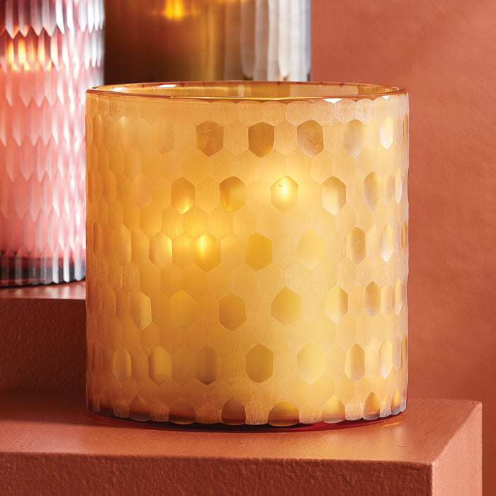 Cream cylindrical lamp with honeycomb cutout pattern, warm amber glow, sits on a pink surface against pink wall.