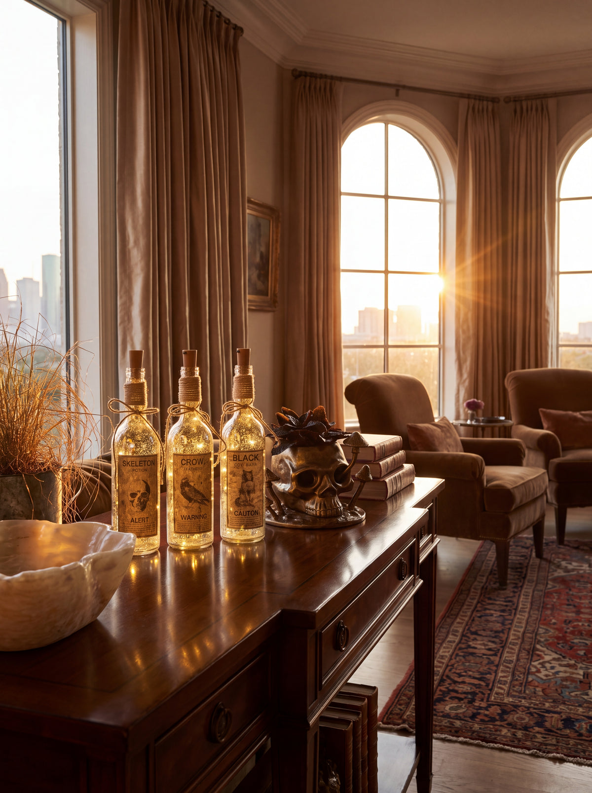 Three decorative clear glass bottles with gold labels and cork stoppers are arranged on a dark wooden console table in a sunlit living room. The room has tall draped curtains, arched windows with warm golden light, upholstered armchairs, a patterned rug, and dried grass decor.