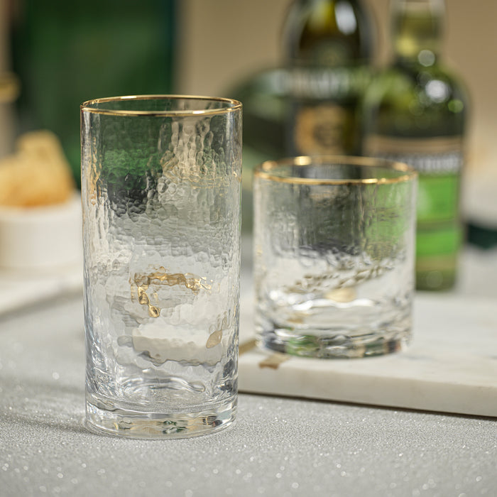Two clear glasses with gold rims on a reflective surface, with bottles in the background.