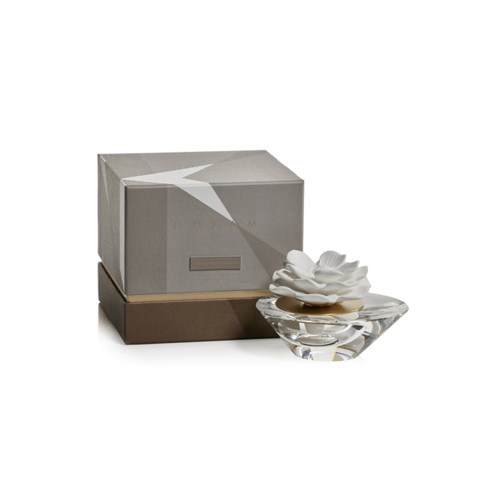 Crystal clear glass diffuser with a white lotus flower top and gold collar, placed beside a gray gift box backdrop.