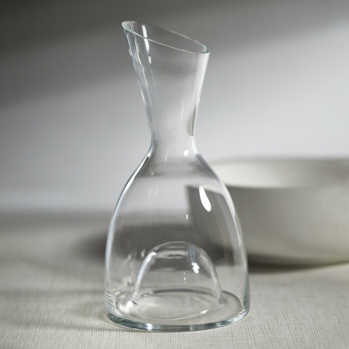 Clear glass wine decanter with an hourglass shape, narrow neck, wide base, set on a light textured surface with a white bowl