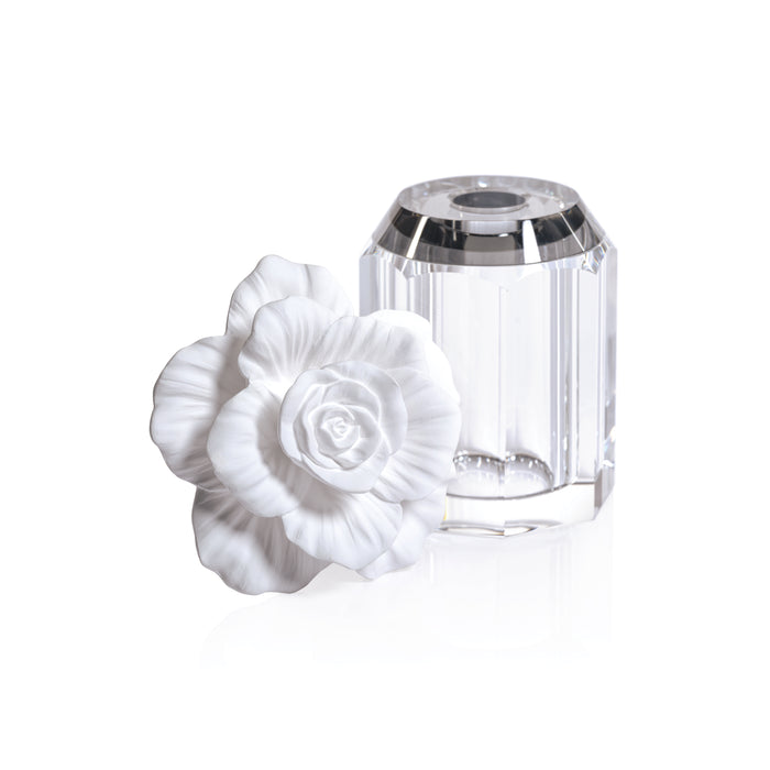 Clear faceted glass diffuser bottle with stopper and a white porcelain camellia flower beside it on a white background.