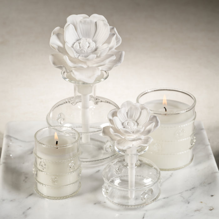 A white porcelain diffuser in the shape of a lotus flower with a glass base, alongside two candle holders with lit candles on a marble surface.