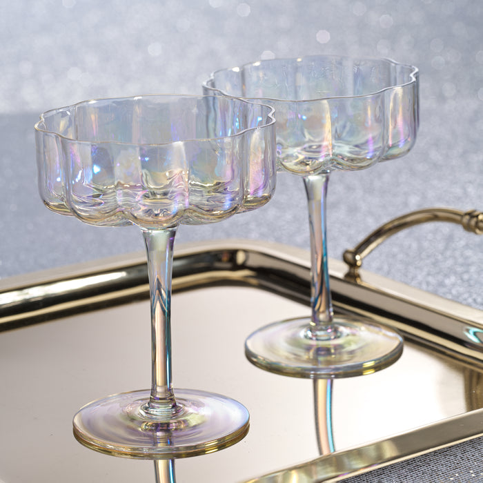 Iridescent glass coupe glasses on a reflective surface with a blurred background
