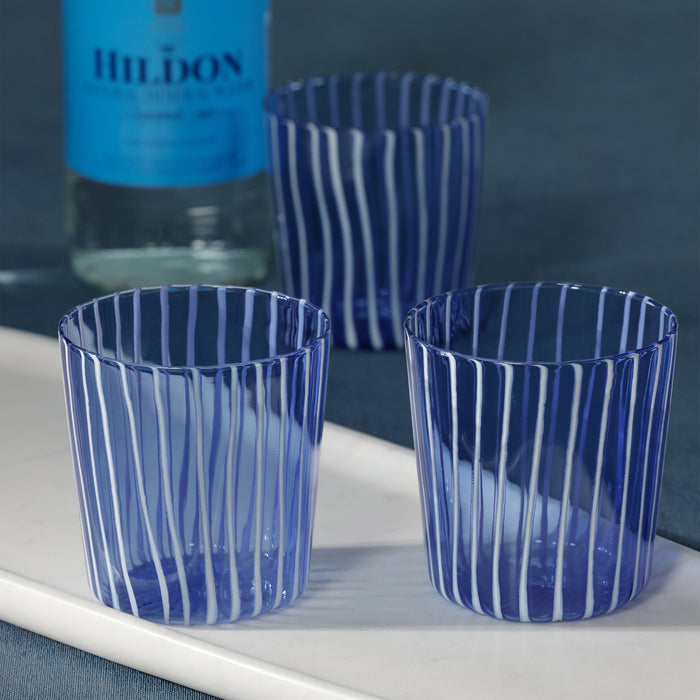 Three blue striped glasses on a white surface with a bottle in the background.