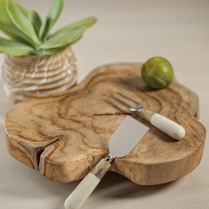 A cheese knife and fork set with marble handles, placed on a wooden cheese board, with a small green fruit and a plant decoration in the background.