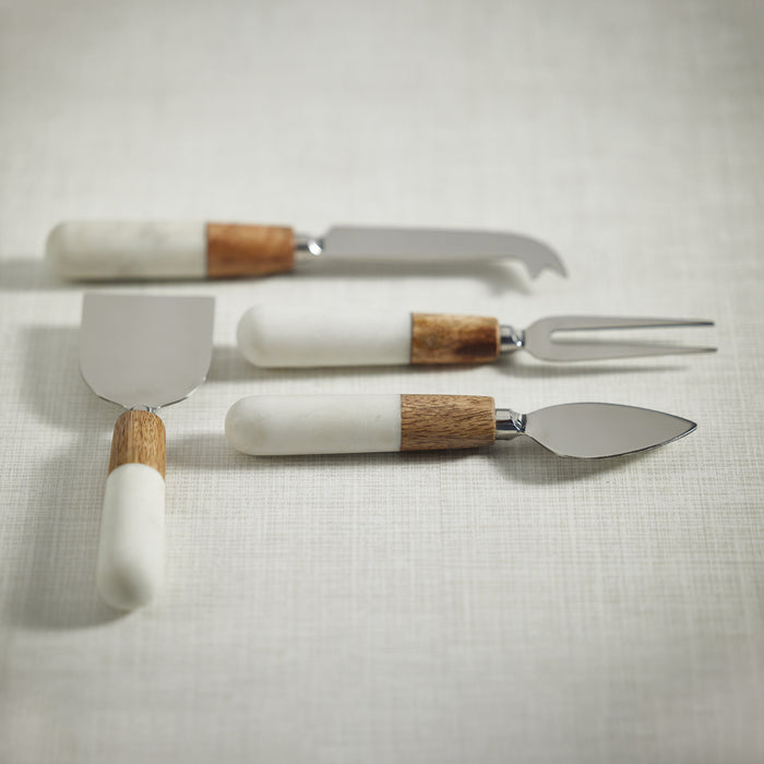 Set of four cheese tools with white marble and wood handles, stainless steel blades, on a light fabric background.