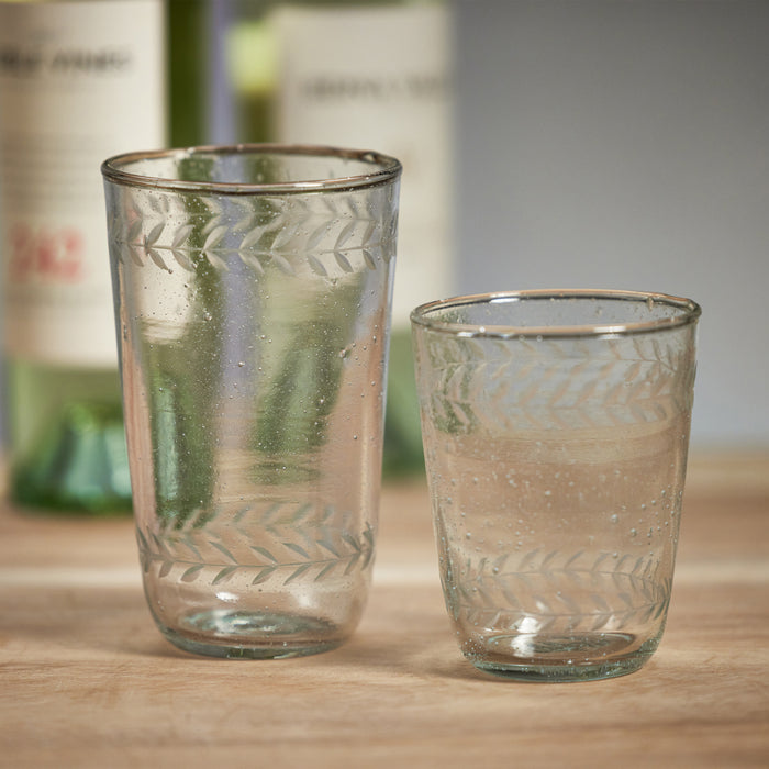 Two etched double old fashioned glasses with leaf pattern, pale blue-green glass, on wooden surface with blurred bottles in