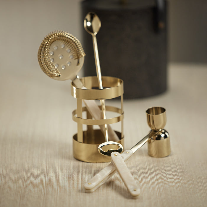 Set of gold cocktail tools including a jigger, strainer, and spoon on a beige surface.
